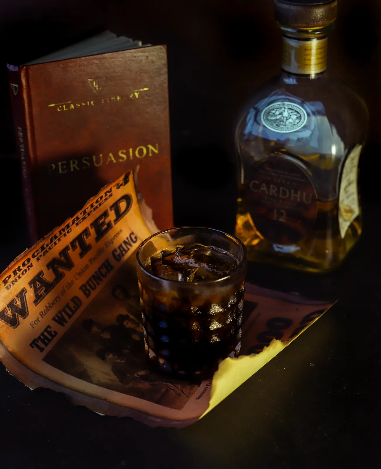 A dark whiskey cocktail on a table beside a bottle and book.